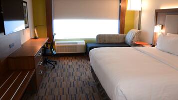A hotel room with a large bed, a desk with a chair, a flat-screen TV, a window with blinds, and a lamp.