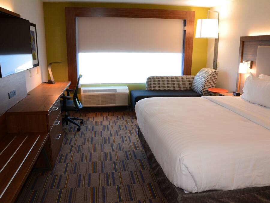 A hotel room with a large bed, a desk with a chair, a flat-screen TV, a window with blinds, and a lamp.
