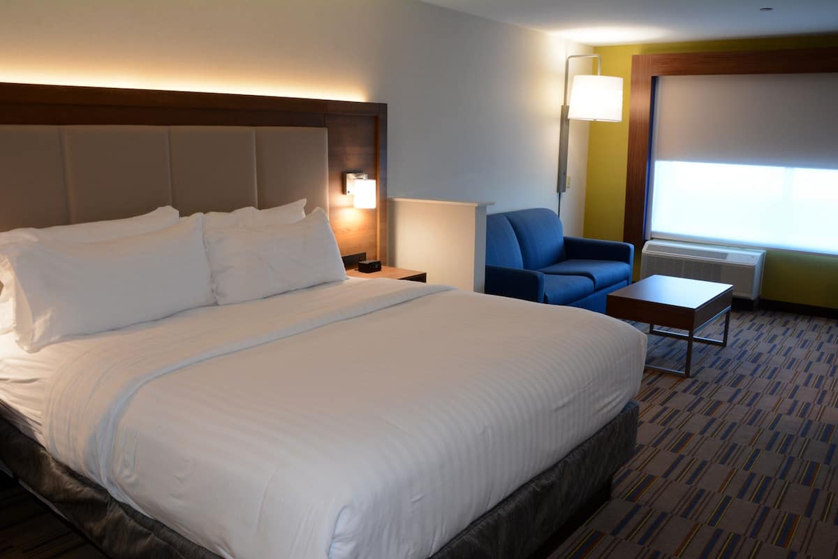 A hotel room with a large bed, a desk, a chair, a lamp, and a window with an air conditioner.