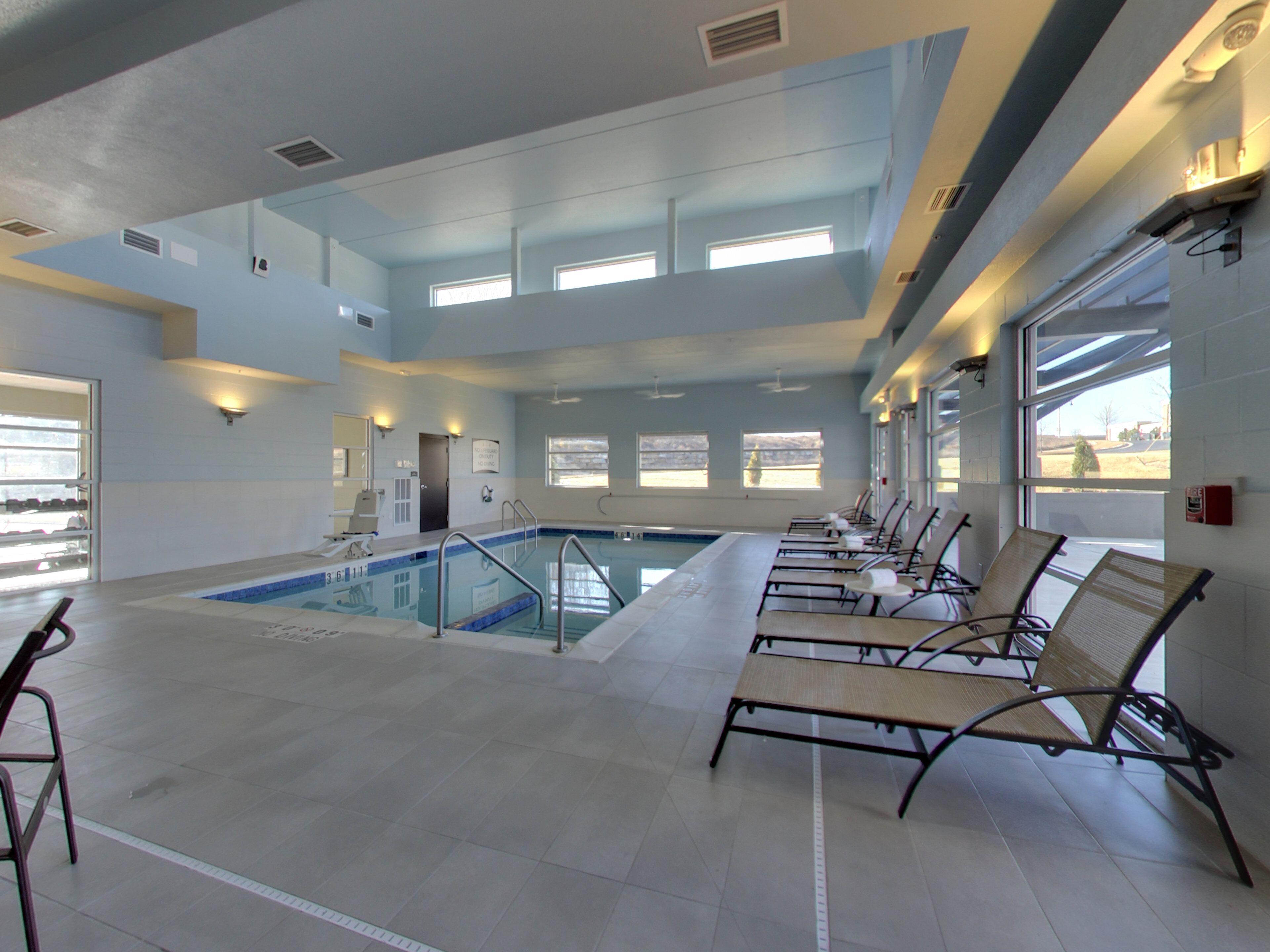 An indoor swimming pool with clear glass walls, a tiled floor, and sunlit windows.