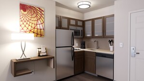 Una cocina compacta con refrigerador, microondas y lavavajillas. Hay un armario de madera con puerta de vidrio, una encimera con cafetera y una repisa de pared con una lámpara y un cuadro enmarcado.