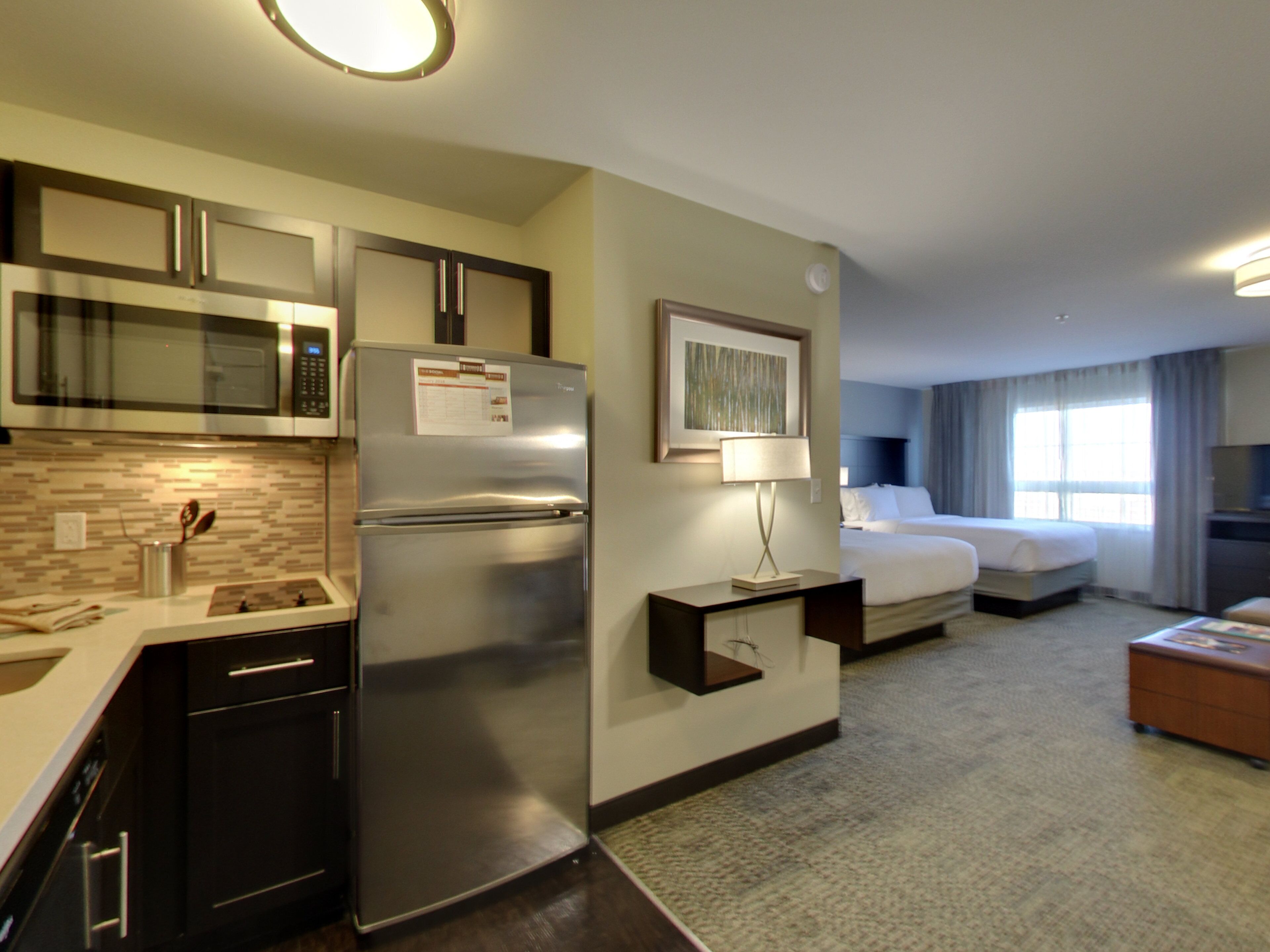 A hotel room with a kitchenette including a refrigerator, microwave, and stove. There is a bed with a headboard, a bedside table with a lamp, and a window with curtains.
