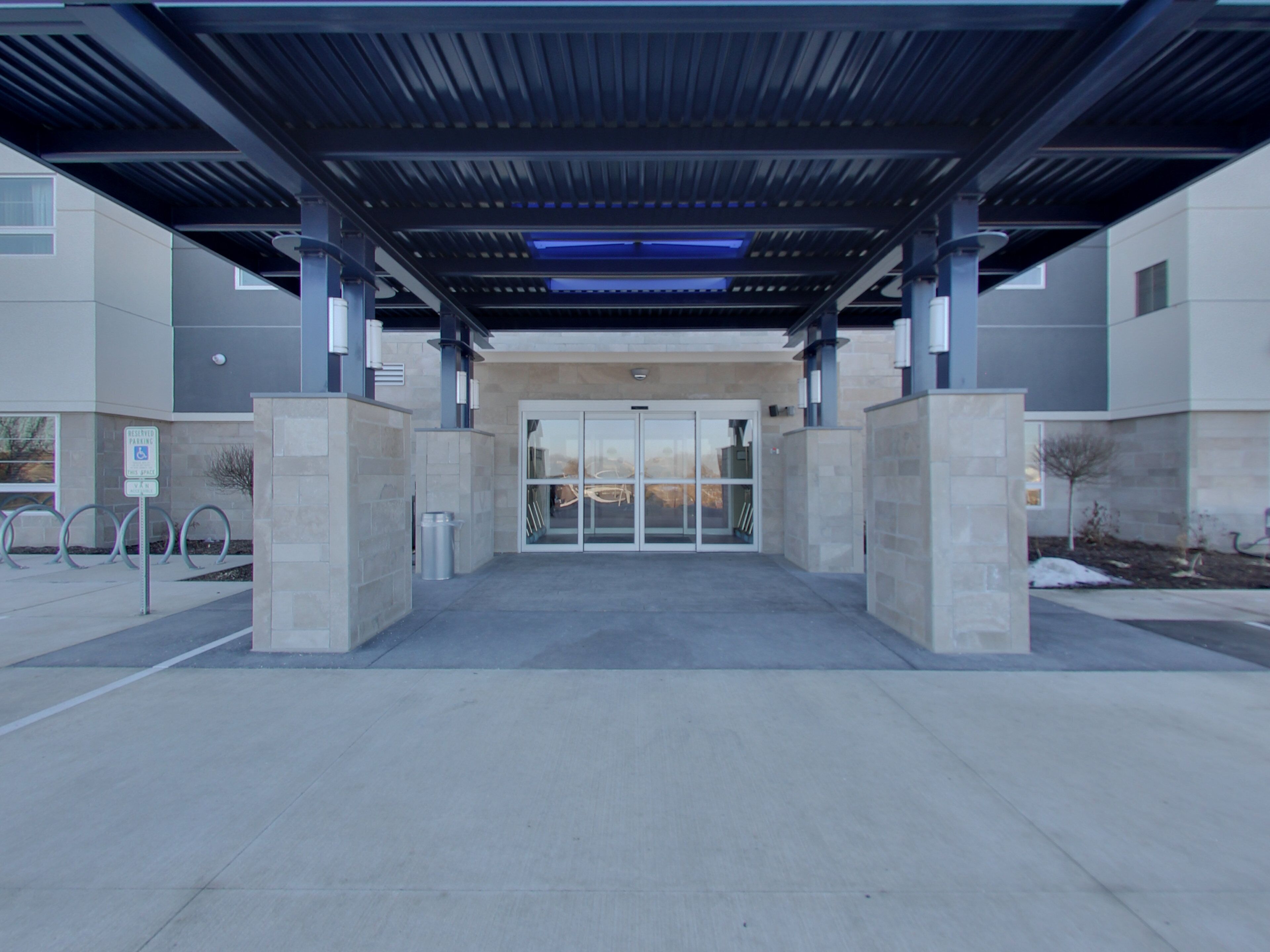 A modern building entrance with a covered walkway, glass doors, and a parking area with designated spaces.