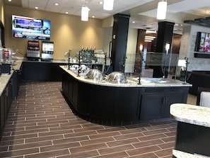 A modern restaurant with a curved counter, a large TV displaying weather updates, and a brick floor. - Staybridge Suites Madison - Fitchburg by IHG (Madison)