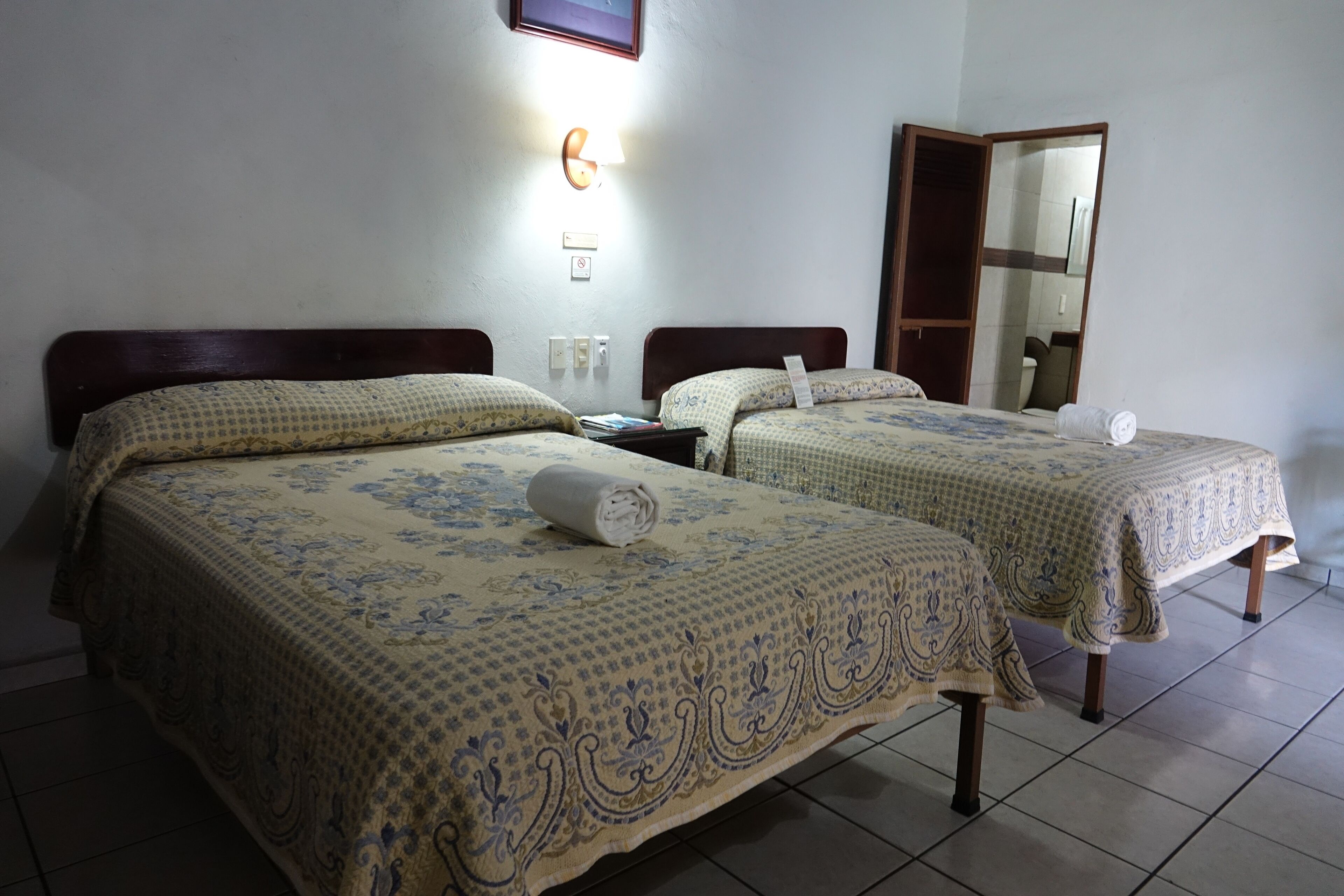 A hotel room with two beds, each with a patterned bedspread, a nightstand, a wall-mounted light, and a mirror.