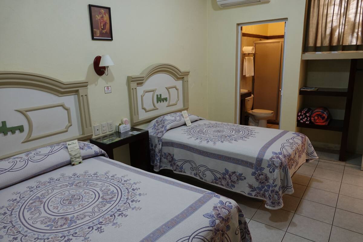 A hotel room with two beds, a nightstand, a wall-mounted lamp, a framed picture, and a bathroom visible through an open door.