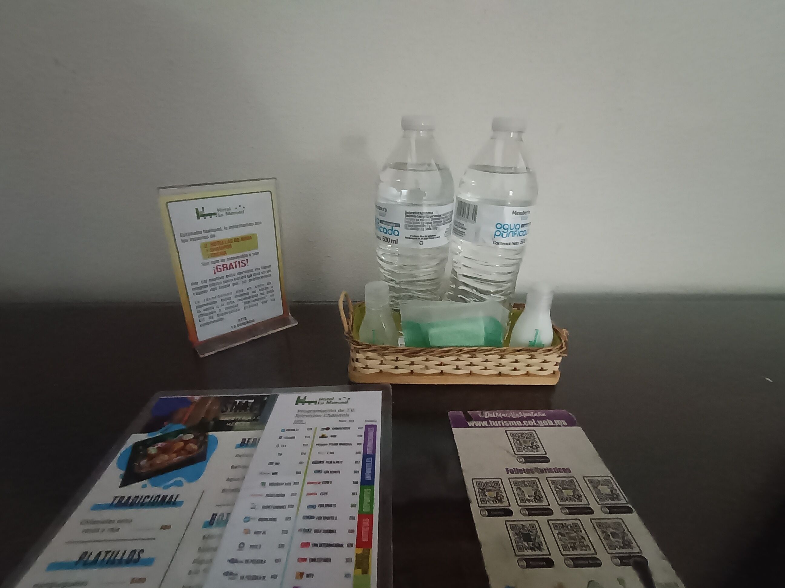 A table with a menu, a QR code for hotel services, and a basket containing water bottles and a green bottle.