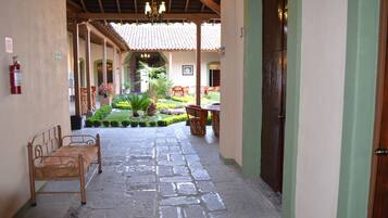 Un corredor cubierto con suelo de piedra, bancos de madera y un pequeño jardín con plantas y sillas.