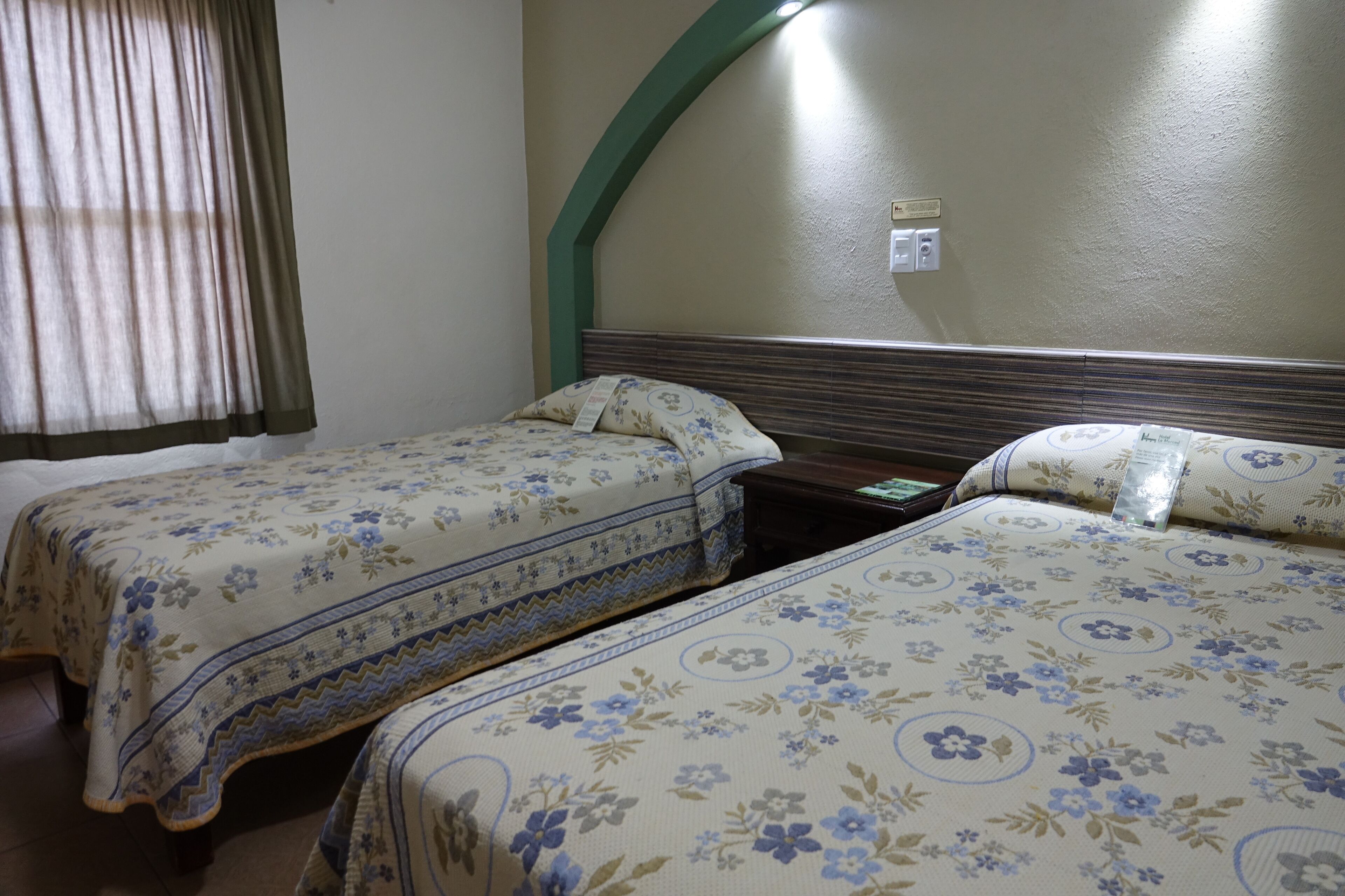A hotel room with two beds, each with floral-patterned bedding, a wooden headboard, and a green arch over the window.