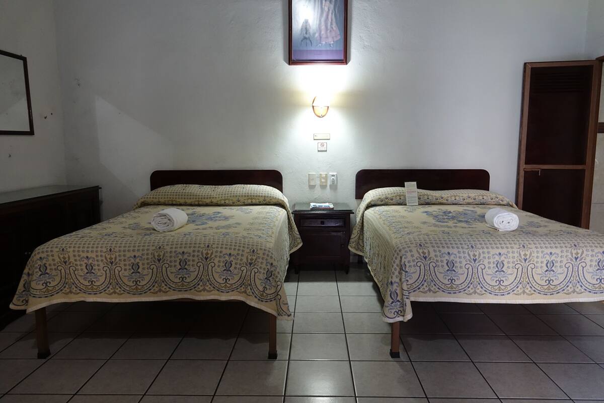 A room with two beds, each with a patterned bedspread and rolled towels. There is a wall-mounted light fixture above the beds and a framed picture on the wall.