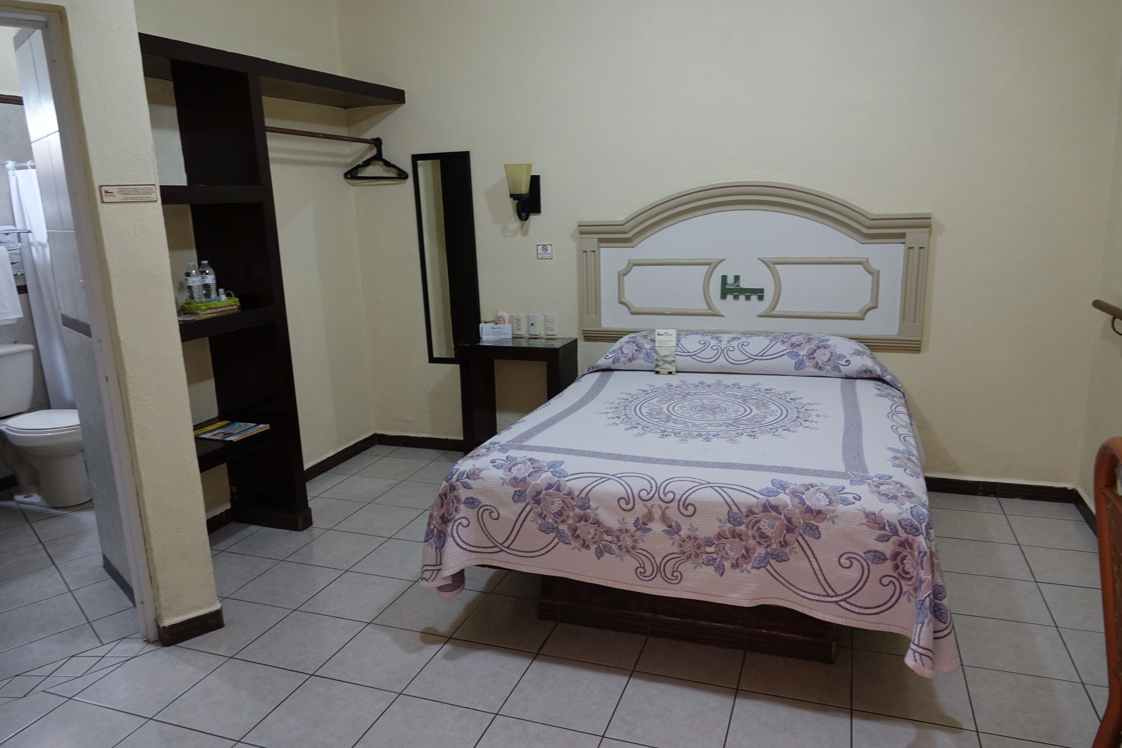 A bedroom with a bed, a nightstand, a mirror, and a shelf with toiletries.
