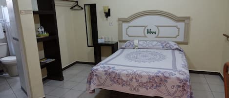 A bedroom with a bed, a nightstand, a mirror, and a shelf with toiletries.
