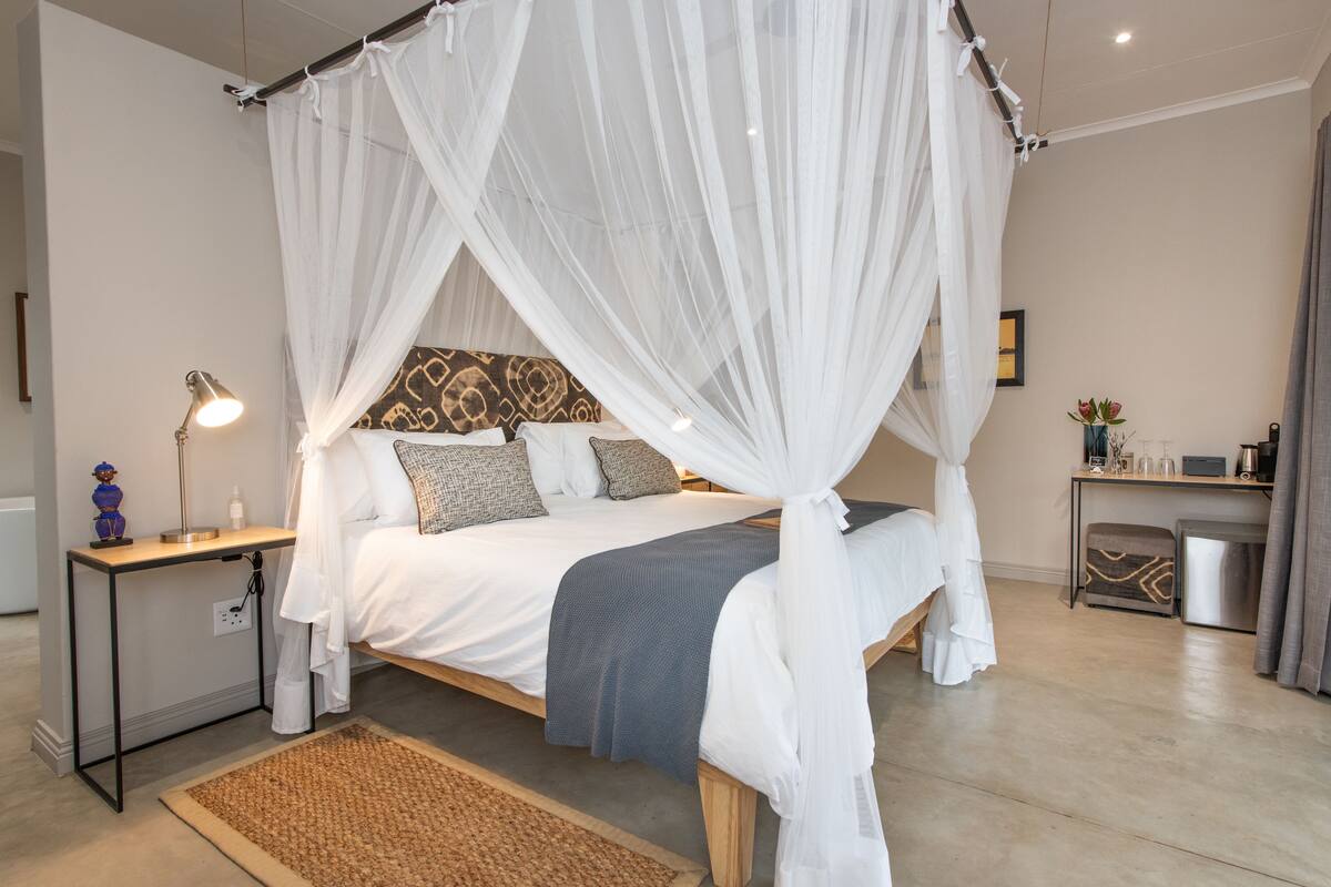 A bedroom with a bed under a canopy, bedside table with lamp, and a small desk with a chair.