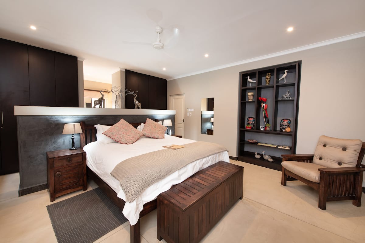 A modern bedroom with a large bed, bedside tables, a bench, a chair, a bookshelf, and a ceiling fan.