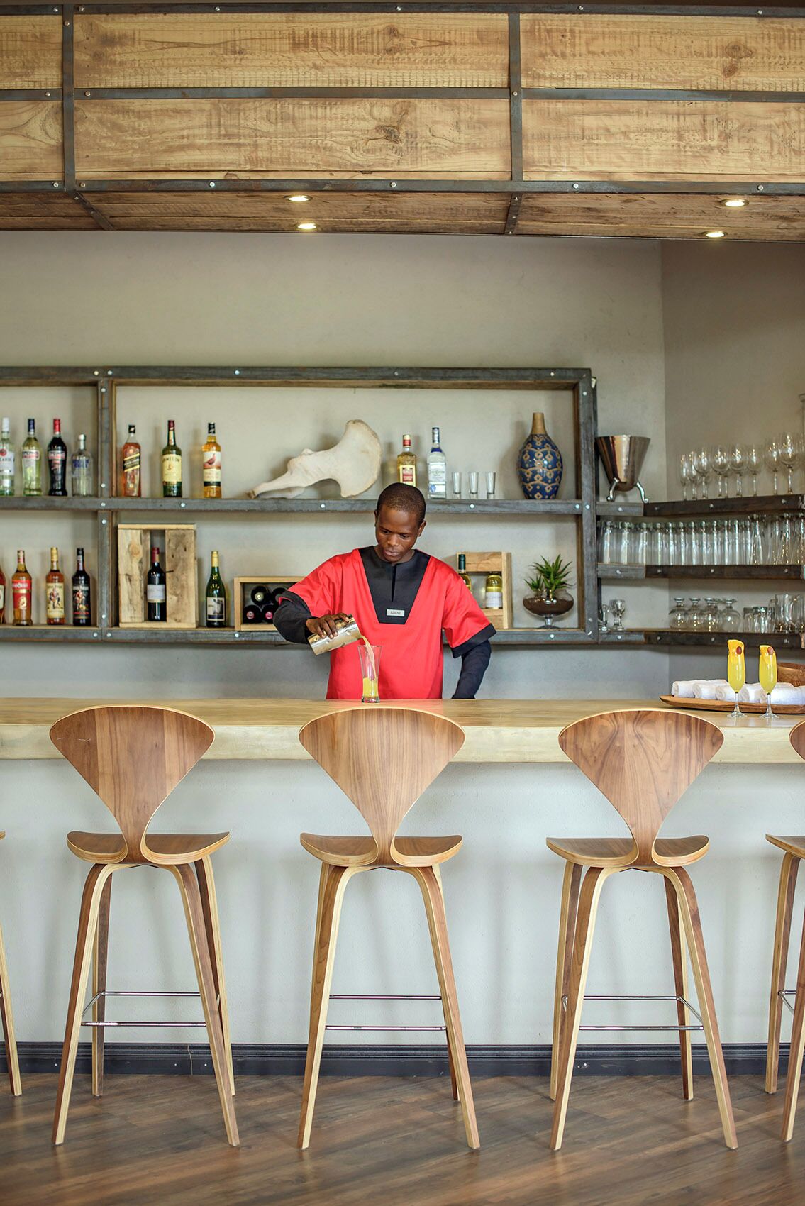 A bar area with wooden chairs, shelves with bottles, and a person pouring a drink.