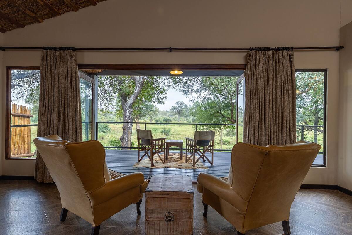 A room with two leather armchairs, a wooden coffee table, and a view of a grassy area with trees through large windows.
