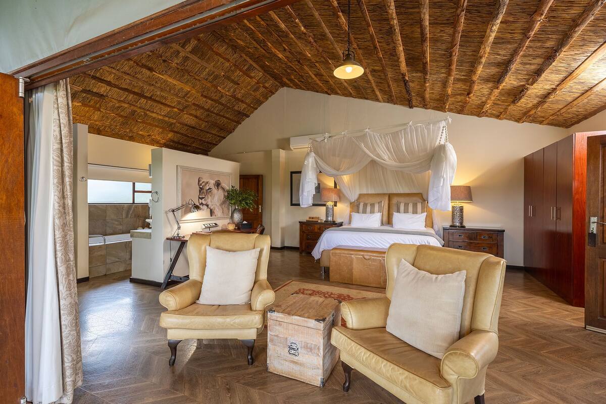 A spacious bedroom with a large bed, a sitting area with two chairs, a wooden ceiling, and a bathroom with a shower and sink.