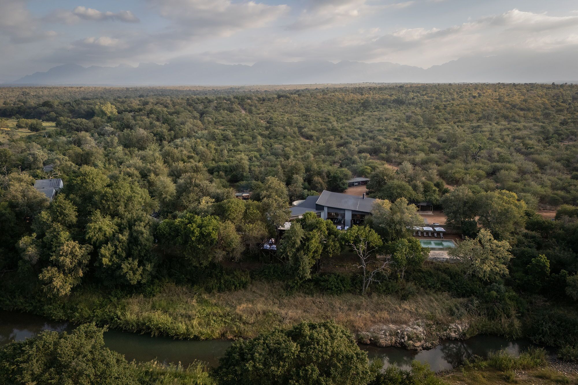 A large house surrounded by dense forest, with a swimming pool and a covered outdoor seating area.