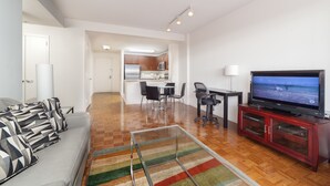 A modern living room with a flat-screen TV, a sofa with patterned cushions, a dining area with chairs, and a kitchen in the background.