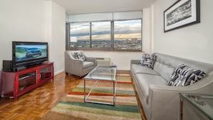 A modern living room with a television, a sofa, a glass coffee table, and a large window offering a city view.