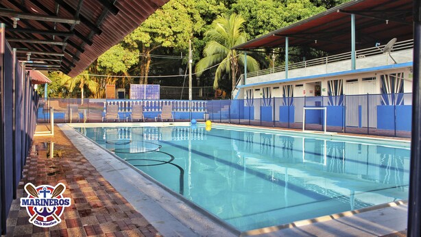 A swimming pool with a covered area, a blue and white building, and a palm tree in the background. - Marineros Club Nautico (Prado)