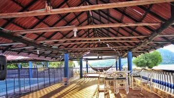 Relax in a scenic outdoor space with a hammock, chairs, and tables overlooking a body of water and mountains.
