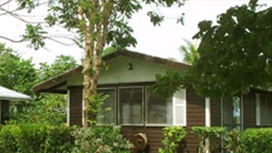 Uma casa de madeira térrea com telhado verde, rodeada por vegetação exuberante e um gramado bem cuidado.