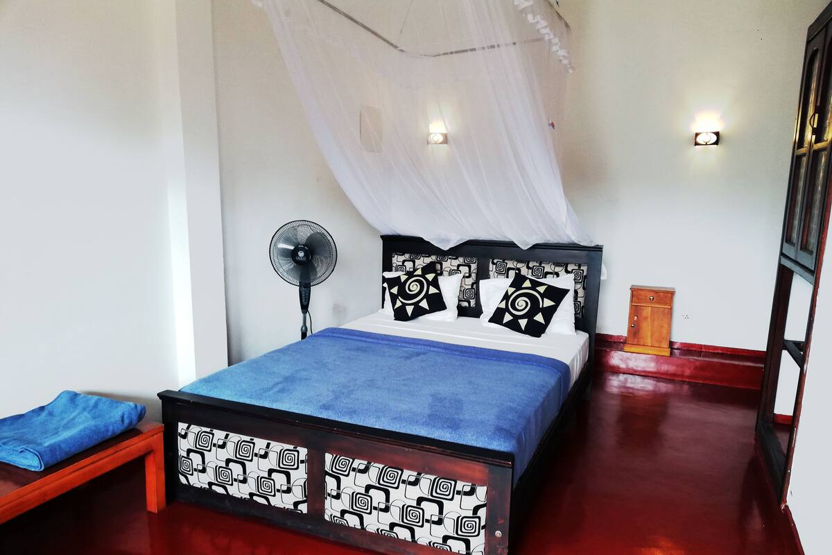 A bed with a patterned headboard, a bedside table, a standing fan, and a nightstand with a lamp.