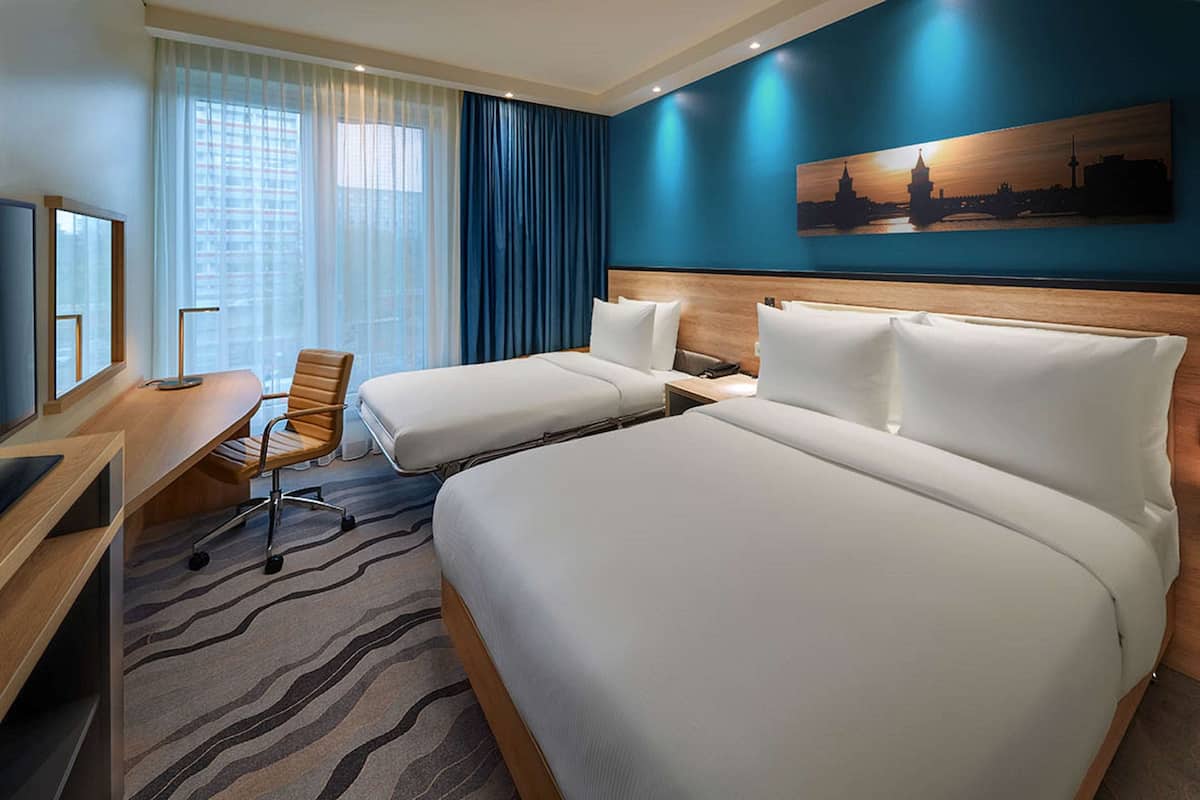 Habitació d'hotel amb dos llits, una taula de treball amb cadira, televisió i una finestra gran amb cortines.