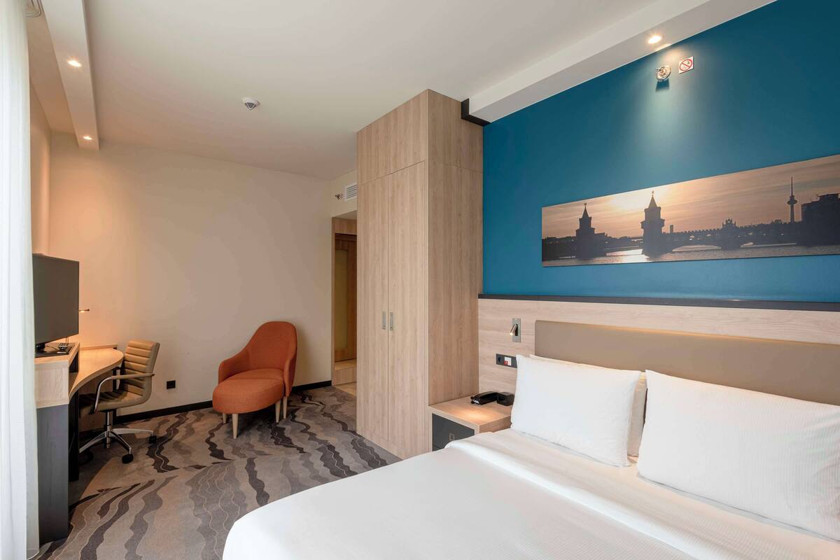 Habitació d'hotel amb un llit, un escriptori, una cadira taronja, un armari i un quadre d'un paisatge urbà a la paret.