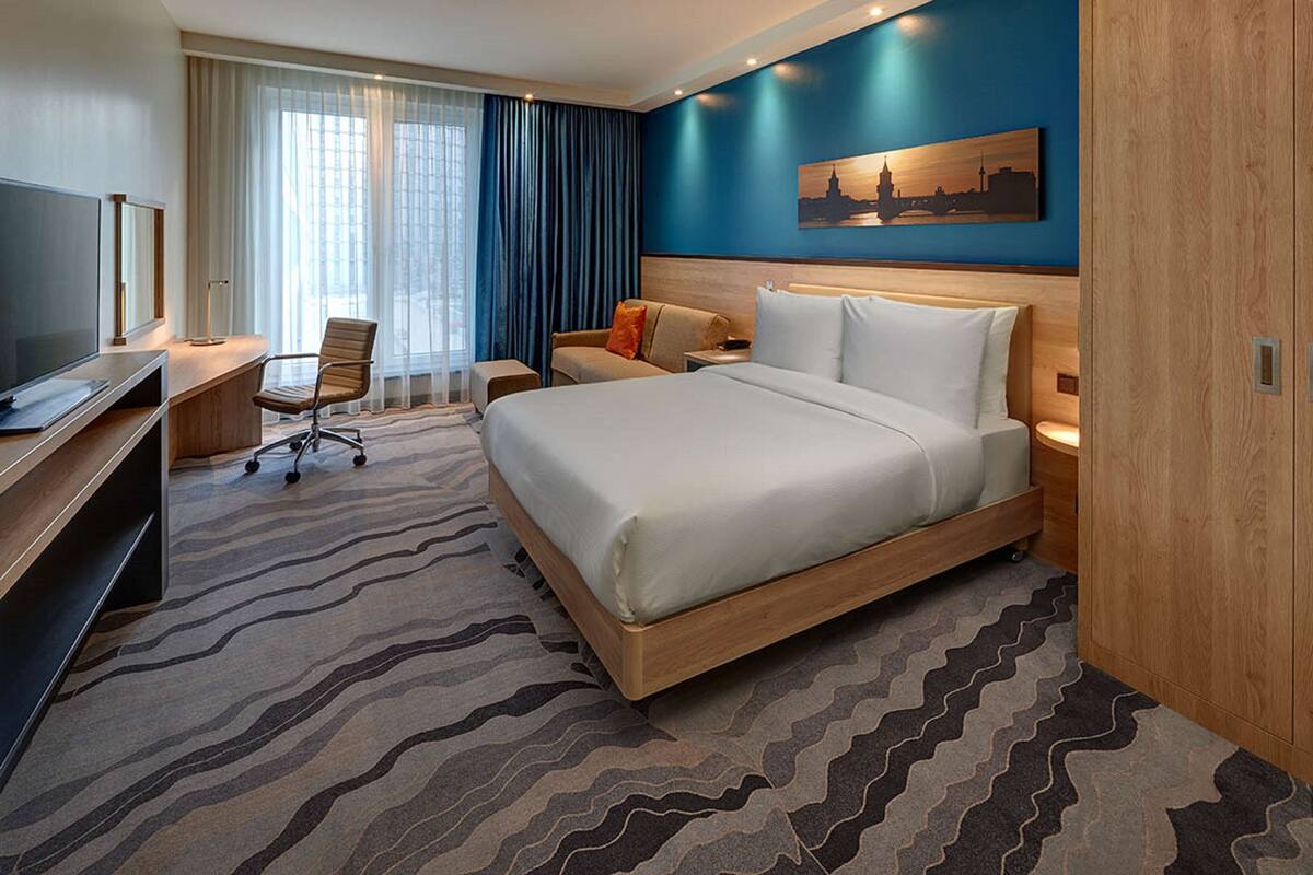 Ein Hotelzimmer mit einem großen Bett, einem Schreibtisch mit Flachbildfernseher, einem Sessel und einer Schreibtischlampe.