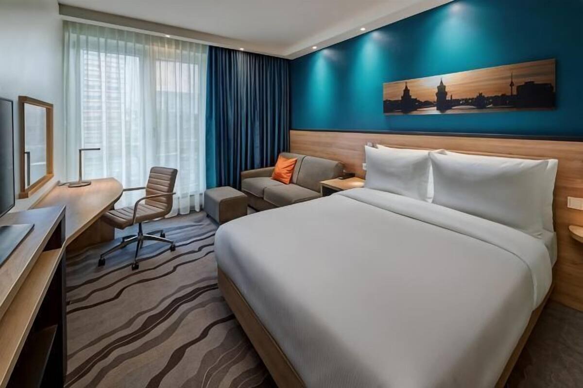 Ein Hotelzimmer mit einem großen Bett, einem Schreibtisch mit Stuhl, einer kleinen Sitzecke und einem Bild an der Wand.