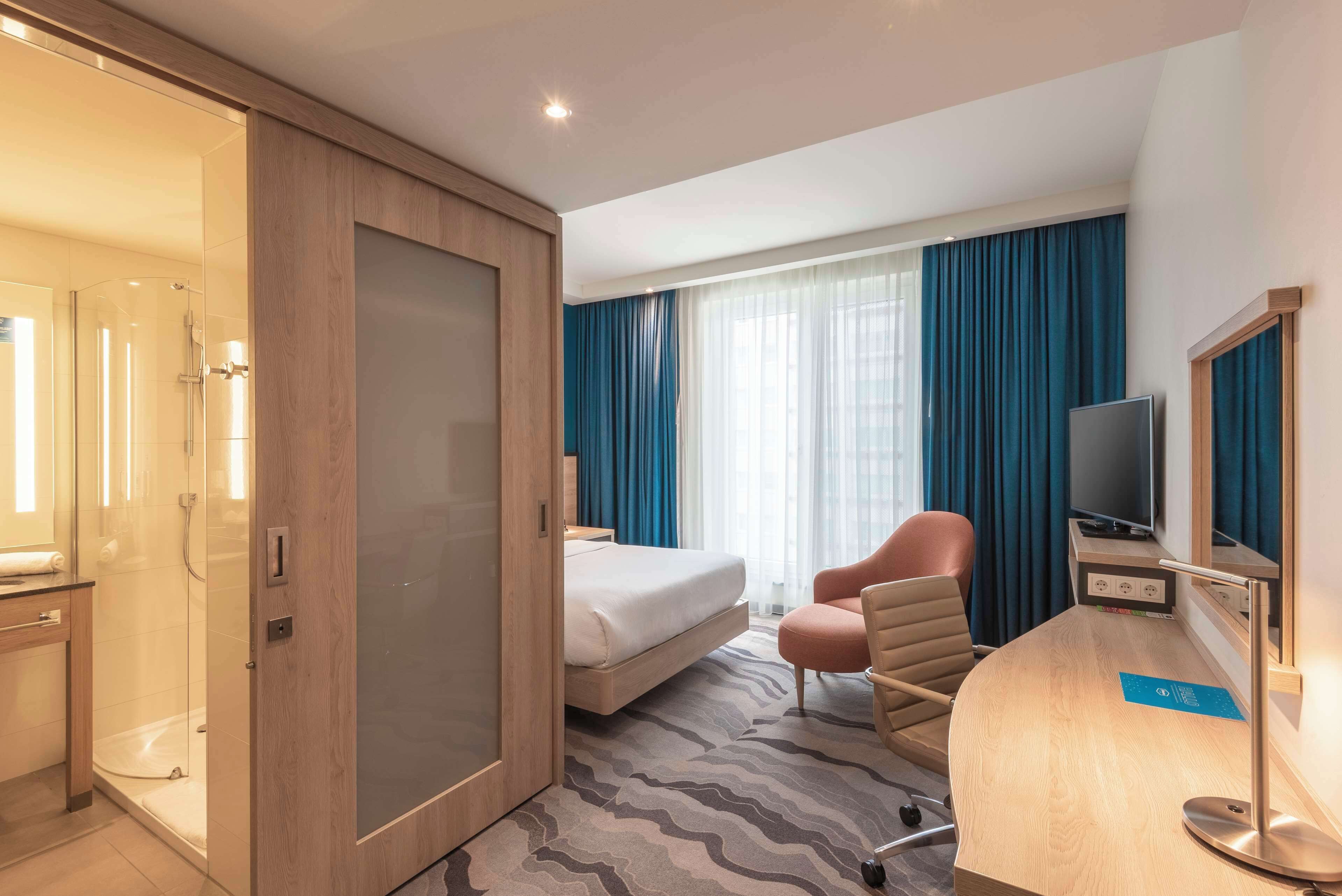 Una camera d'albergo con un letto, una scrivania con un computer, una sedia, una TV e un angolo doccia.