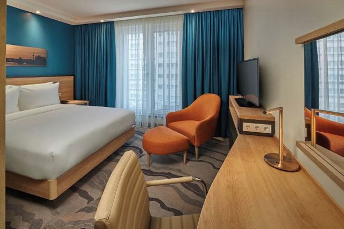 A hotel room with a bed, a desk with a computer, an orange chair, and a large mirror.