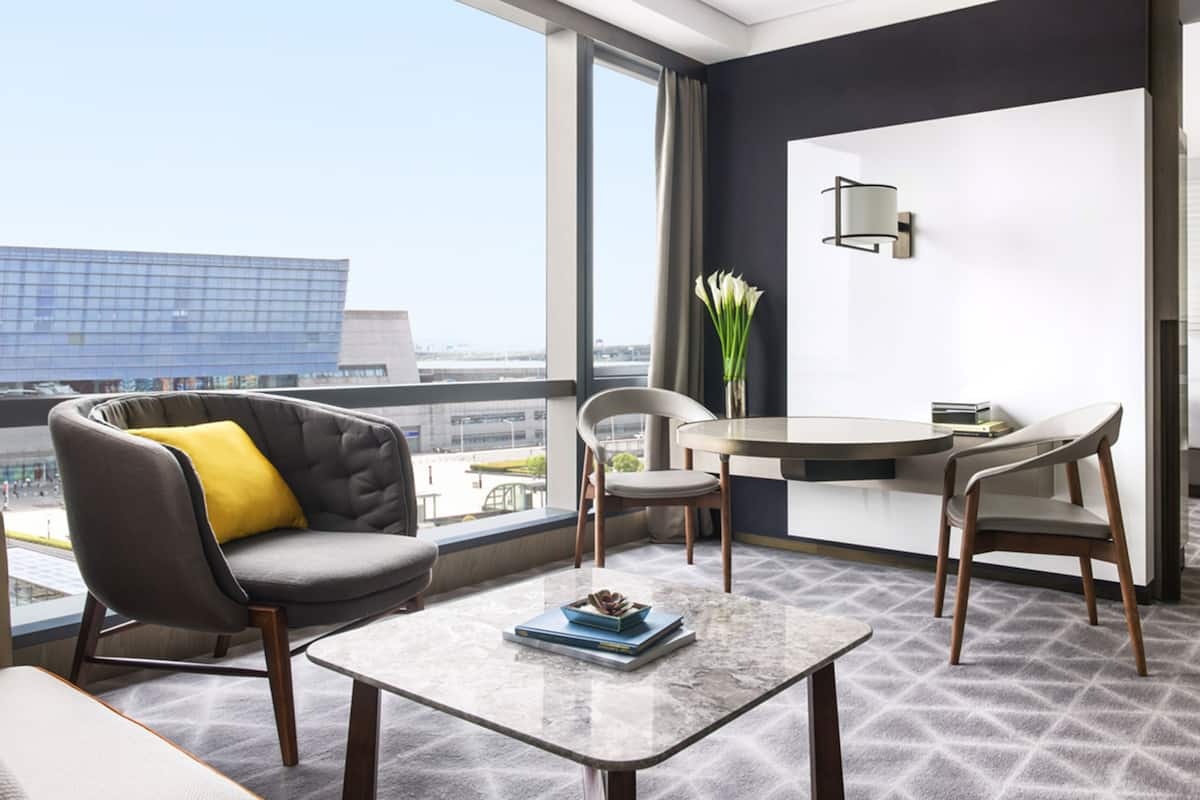 Una habitación de hotel moderna con un balcón, una mesa redonda, dos sillas, un sofá con un cojín amarillo, un florero con flores y vistas a la ciudad.