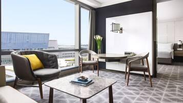 A modern hotel room with a balcony, a round table, two chairs, a sofa with a yellow pillow, a vase with flowers, and a view of the cityscape.