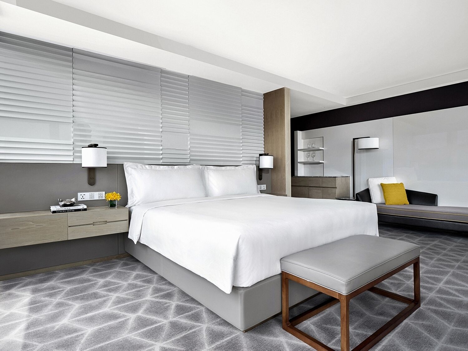Ein modernes Hotelzimmer mit einem großen Bett, Nachttischlampen, einem Nachttisch mit Blumenvase und einer Bank.