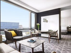 Una habitación de hotel moderna con un ventanal grande, una mesa redonda para comer, un sofá con un cojín amarillo y una cama con una mesita de noche.
