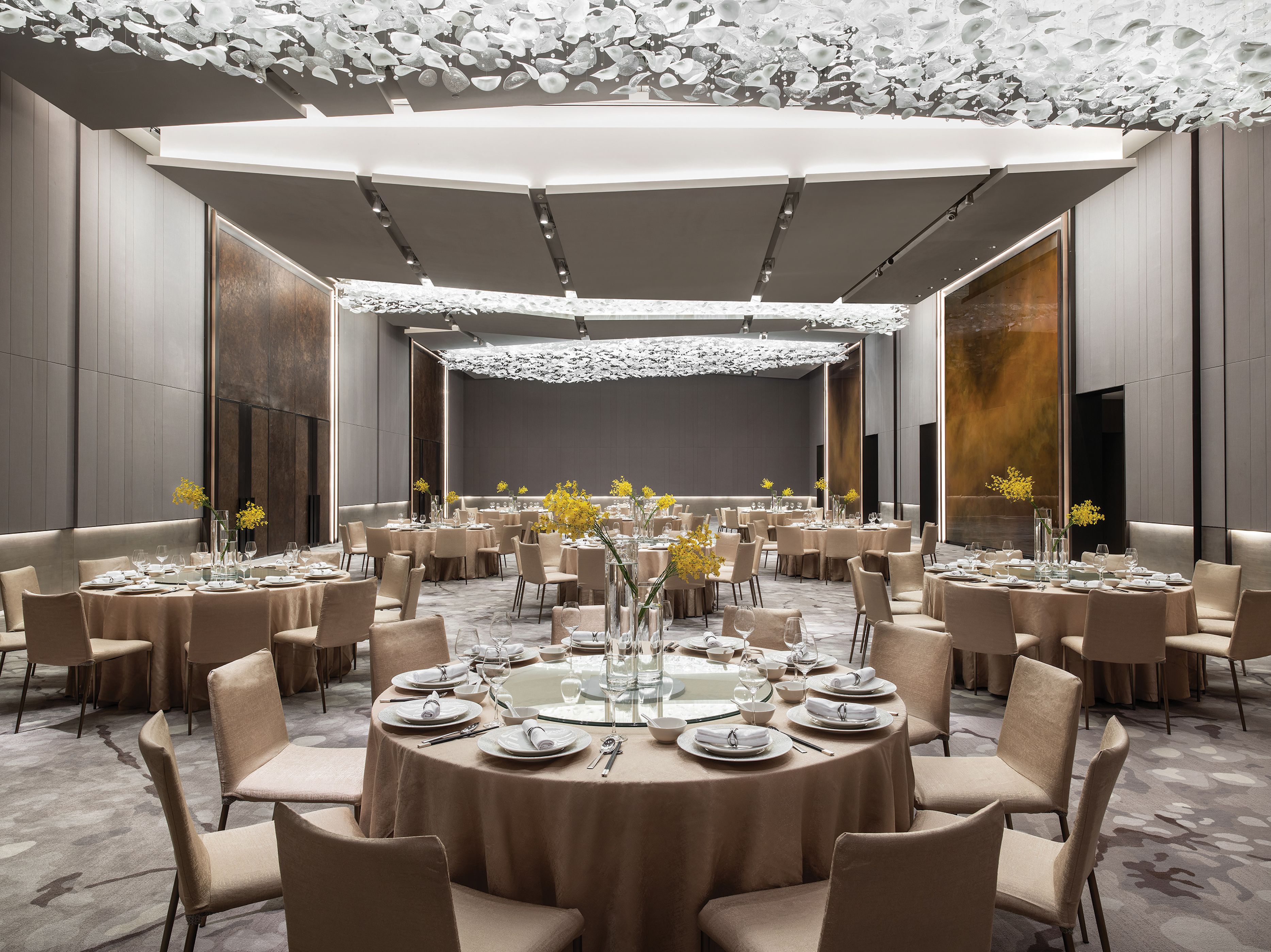 Un amplio y elegantemente decorado salón de banquetes con mesas redondas dispuestas para cenar, con un arreglo floral central y un techo alto con una lámpara única.