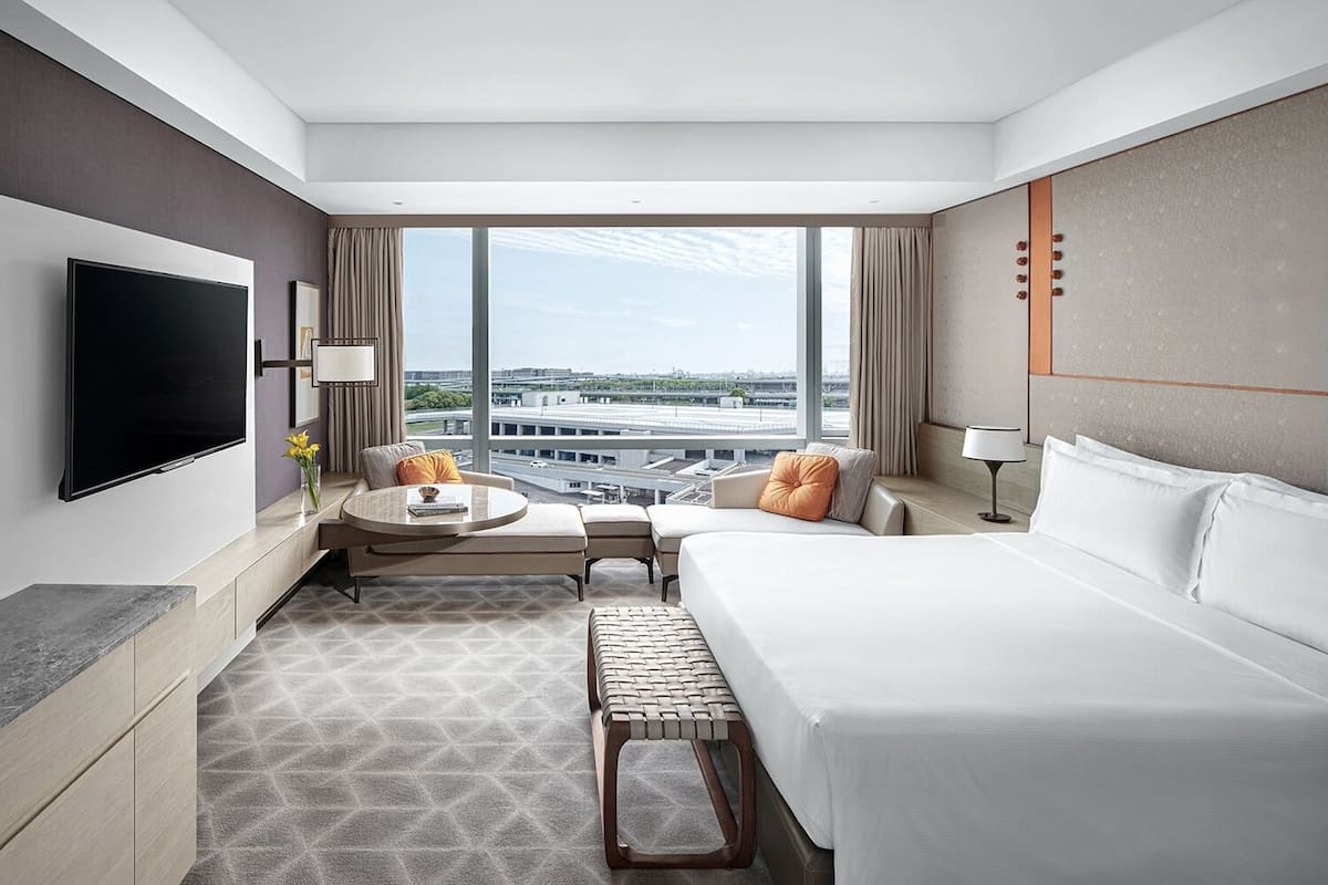 Habitación de hotel con cama, televisión, escritorio y una ventana grande con vista a la ciudad.