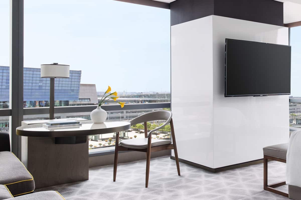 Habitación de hotel con cama, escritorio con silla, televisor montado en la pared y un ventanal con vista a la ciudad.