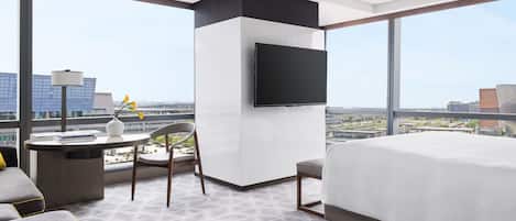 A hotel room with a bed, a desk with a chair, a TV mounted on the wall, and a large window offering a city view.