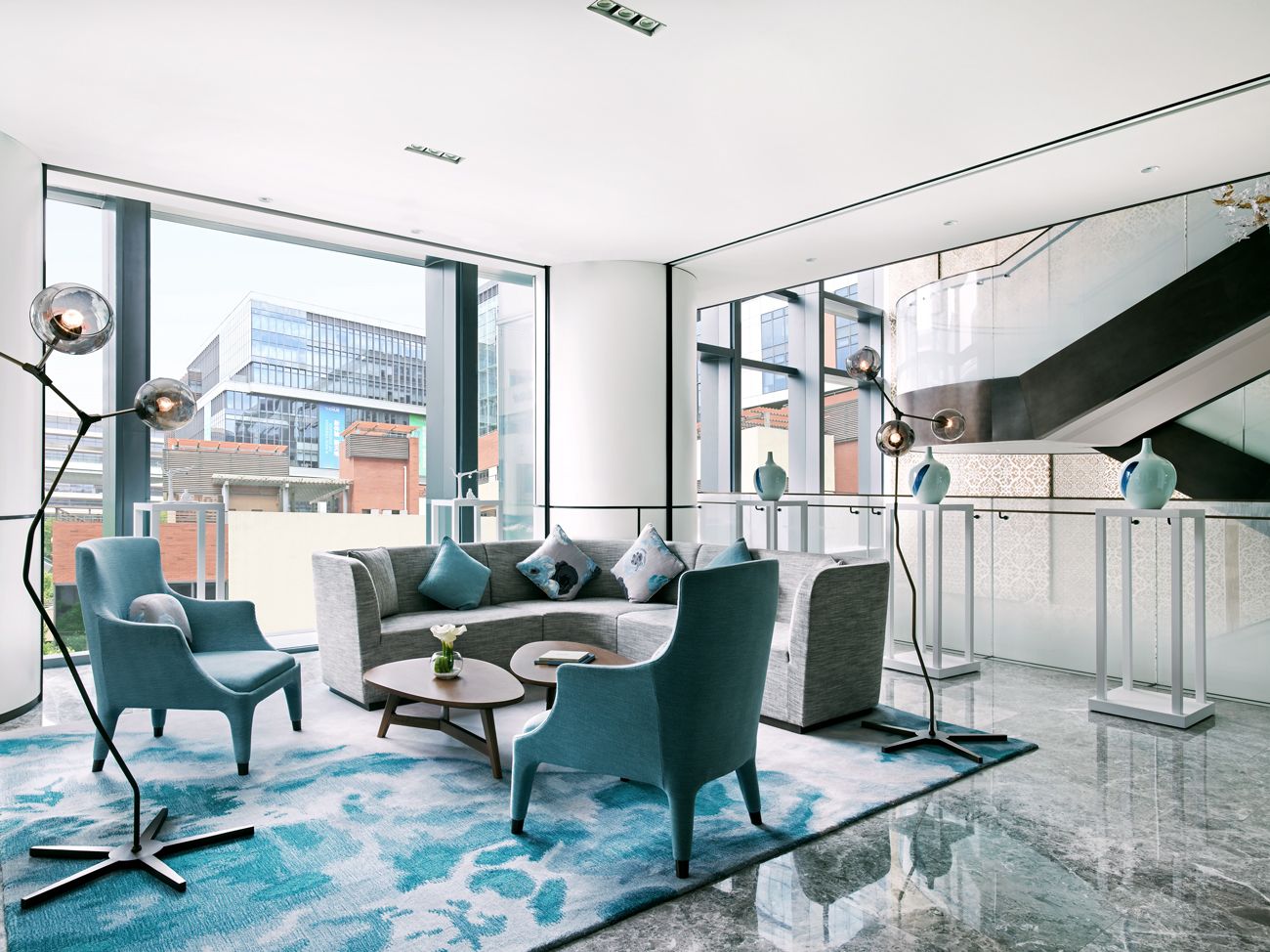 A modern living room with a sofa set, armchairs, and a coffee table. There are large windows offering a view of the cityscape.
