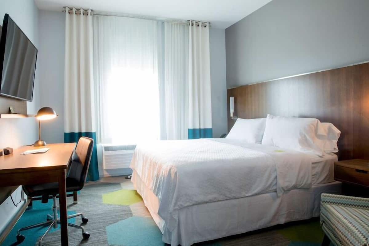 A hotel room with a bed, a desk with a chair, a TV, and a window with curtains.