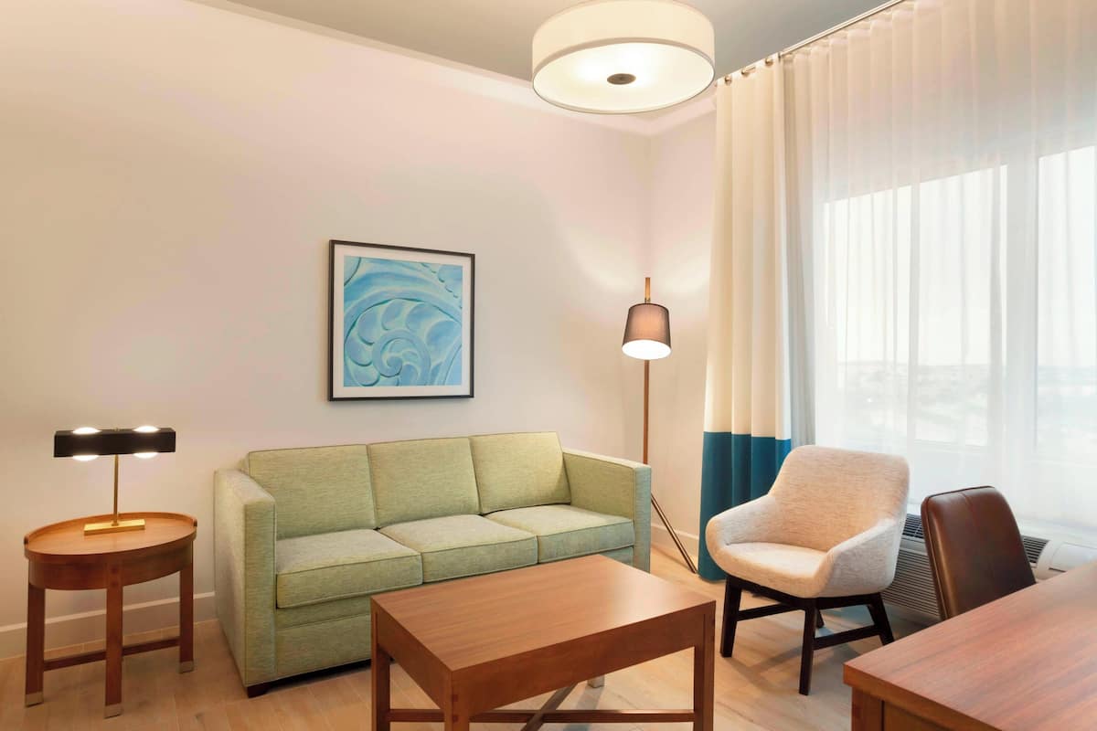 A modern hotel room with a green sofa, a wooden coffee table, a chair, a lamp, and a painting on the wall.