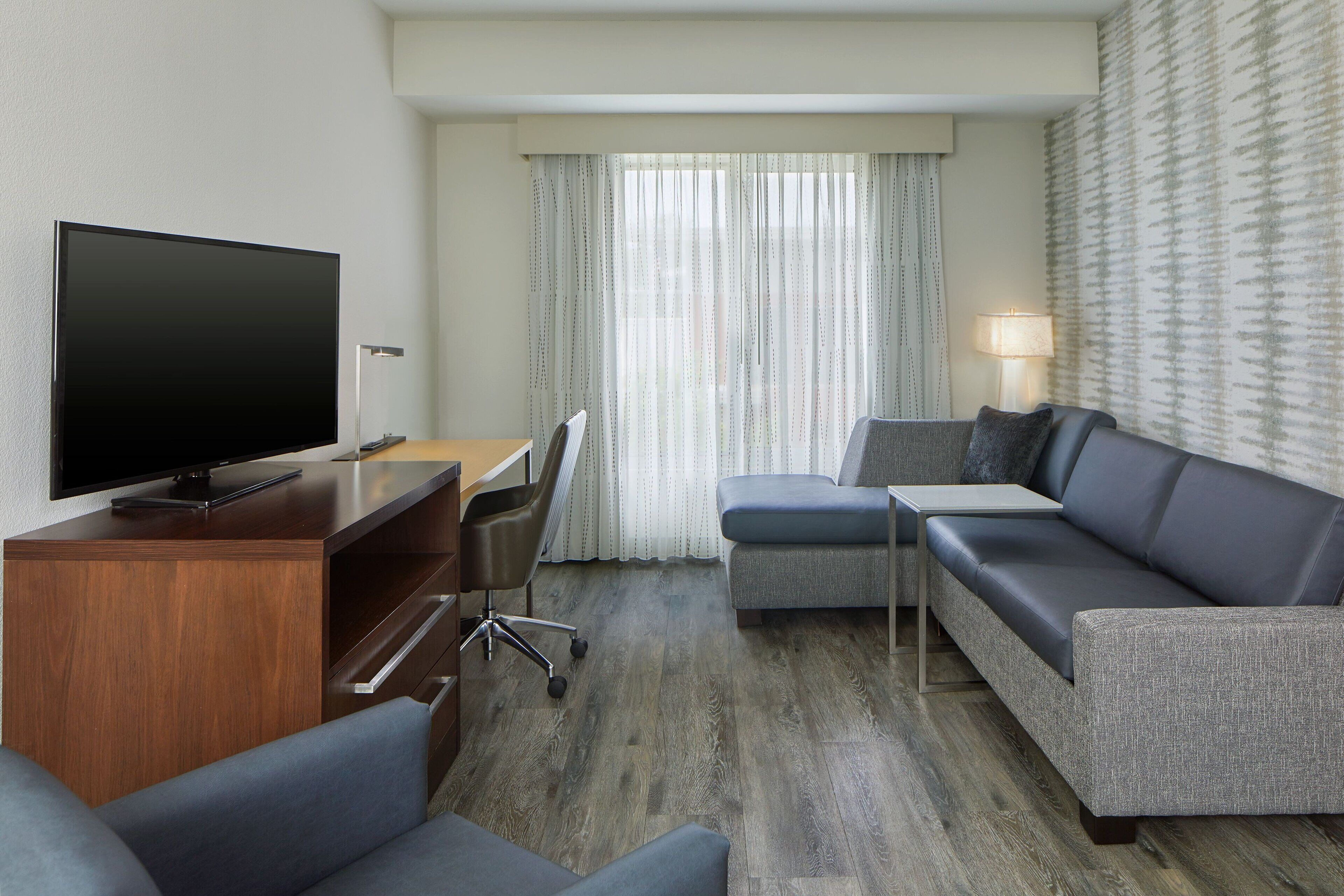 A modern hotel room with a flat-screen TV, a wooden desk, a grey sofa, and a small table.