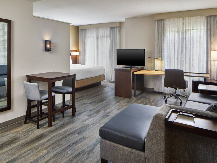 A hotel room with a bed, a desk with a TV, a sofa, and a dining area with chairs.