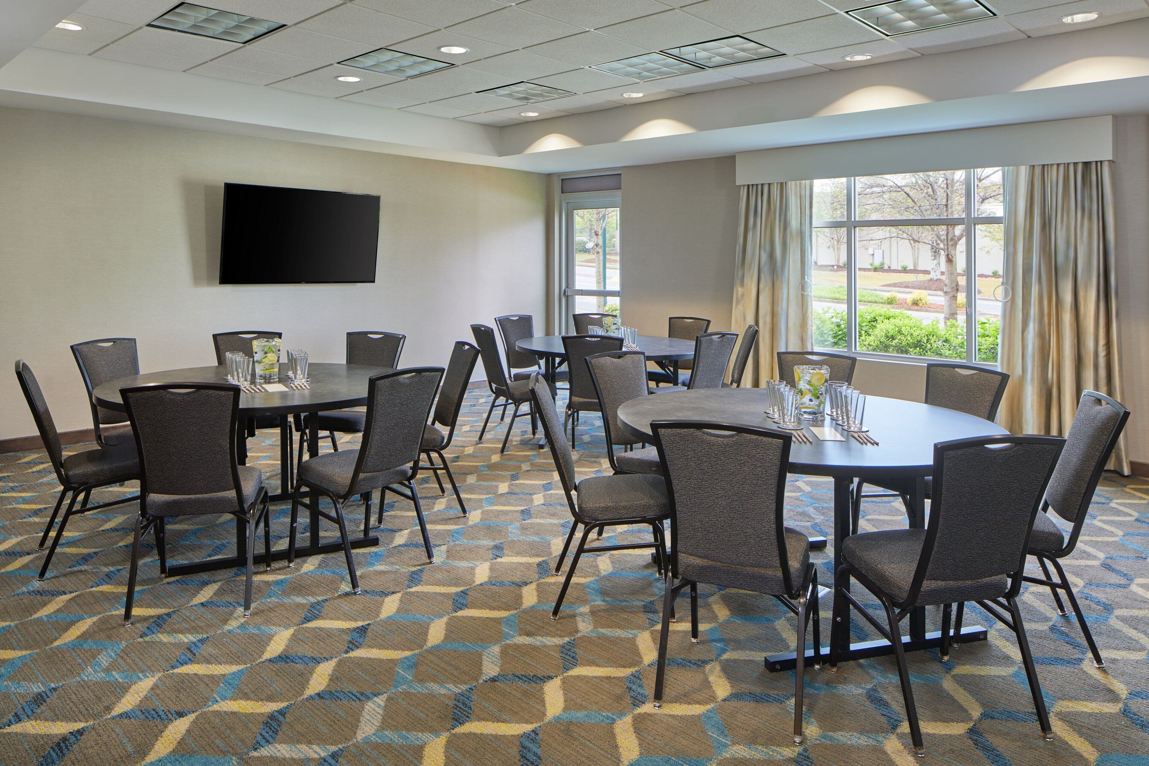 A conference room with multiple tables and chairs, a flat-screen TV on the wall, and large windows with curtains.