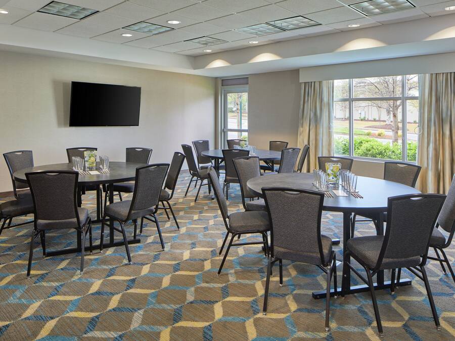 A conference room with multiple tables and chairs, a flat-screen TV on the wall, and large windows with curtains.