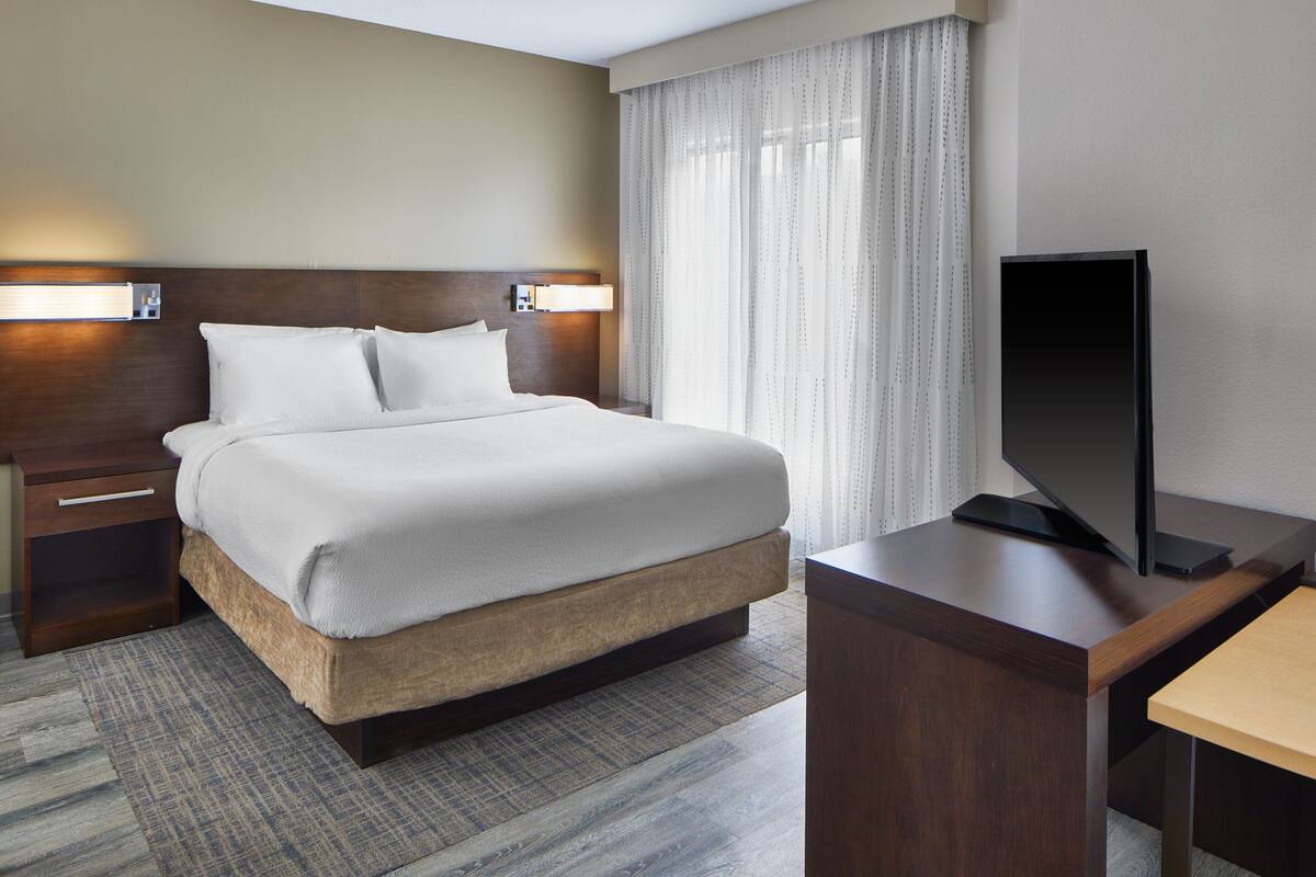 A hotel room with a large bed, a flat-screen TV on a wooden stand, a nightstand, and a window with sheer curtains.